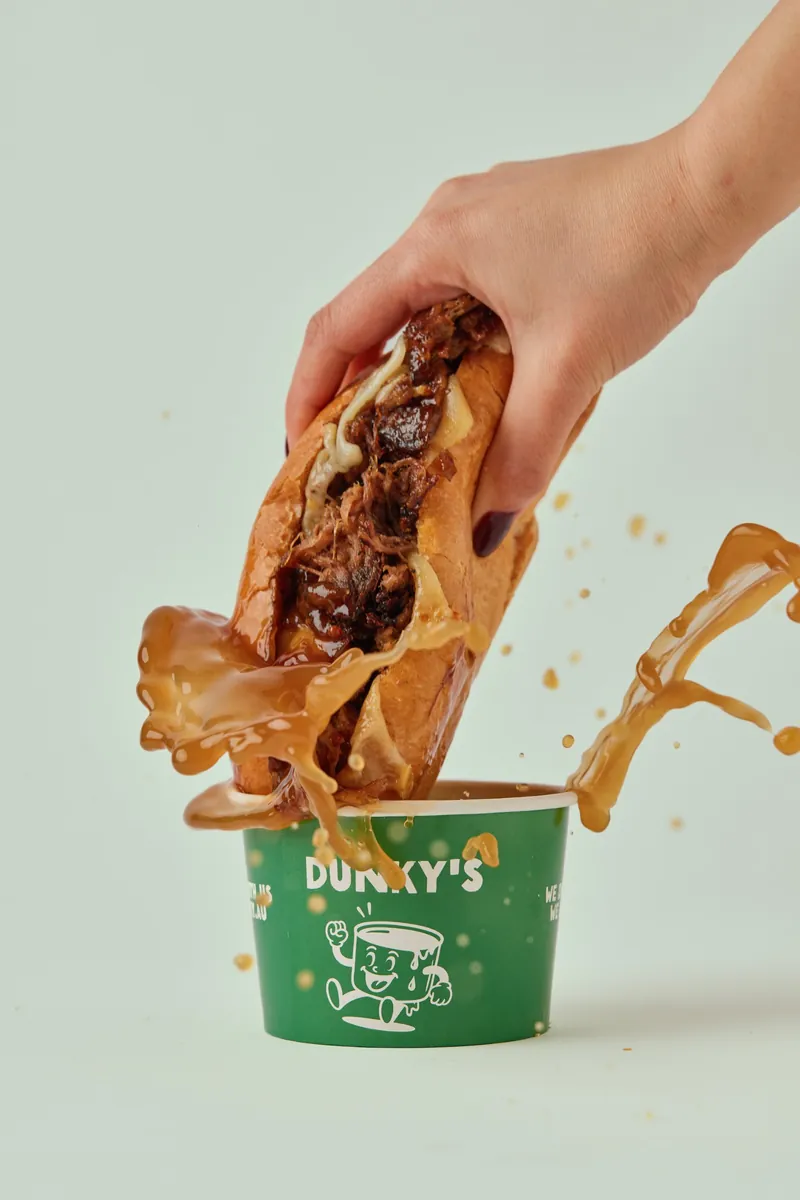 The OG Brisket roll being dunked into a Dunky's cup of beef jus, splashing over the sides